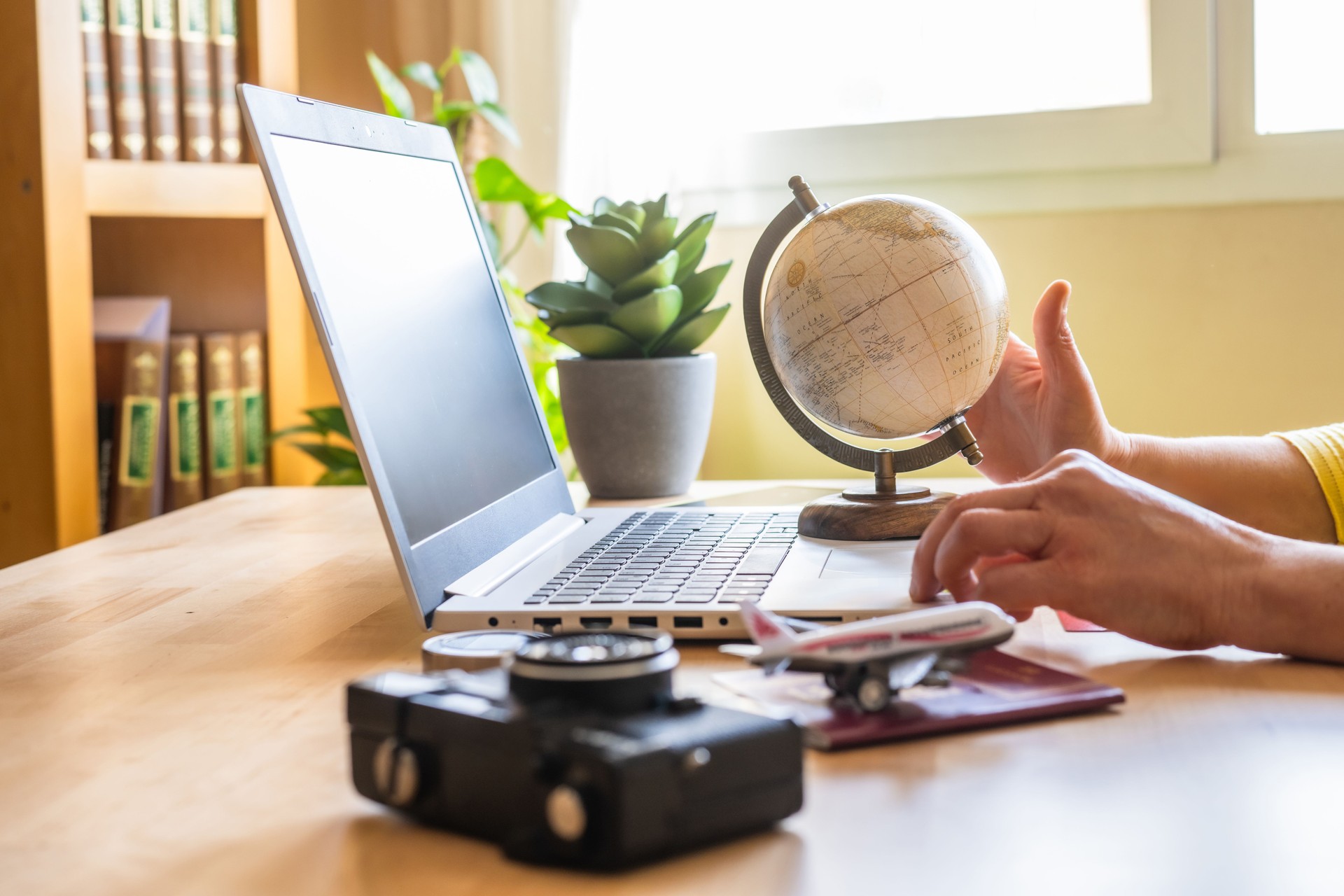 Woman planning a trip with a laptop, touching a globe while booking a vacation online. Airplane model, passport, camera, and compass arranged on the desk, creating a travel atmosphere